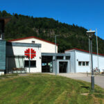 Außenansicht der Turnhalle. Der linke Teil hat eine türkisfarbene Holzverkleidung. Rechts daneben ist eine Sitzecke zu sehen und mittig im Bild ist der Eingang. Auf der rechten Seite ist ein Anbau sichtbar, vor dem Fahrräder und E-Scooter angeschlossen sind.