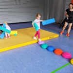 Die Szene zeigt drei Kinder in einer Turnhalle, die mit verschiedenen Dingen spielen. Im Hintergrund steht eine Frau mit einem Baby vor dem Bauch, die zuschaut.