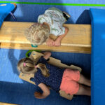 Zwei Mädchen spielen mit einem Rollbrett, zwei Bänken und zwei Turnmatten. Ein Mädchen liegt auf dem Rollbrett. Das andere beugt über eine Bank und schaut zu.