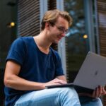 Mann mittleren Alters sitzt im Außenbereich und schaut lächelnd auf den aufgeklappten Laptop-Bildschirm.