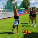 Ein Mädchen des TSV Aistaig macht Handstandlauf auf dem Sportplatz. Zwei Mädchen schauen zu.