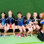 Eine Gruppe Mädchen in abgestimmten Turnanzügen sitzt mit einem riesigen Teddybär in blauem Trikot auf einer Turnhallenbank