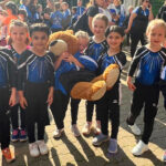 Eine Gruppe Mädchen in einheitlichen Gymnastikanzügen und Turnschuhen steht auf einem Platz. Sie gucken in die Kamera, einige Menschen stehen um sie herum.