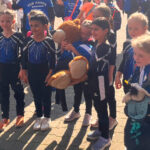 Eine Gruppe Mädchen in einheitlichen Gymnastikanzügen und Turnschuhen steht auf einem Platz. Sie gucken in unterschiedliche Richtungen, einige Menschen stehen um sie herum.