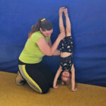 Ein Mädchen macht Handstand gegen eine blaue Weichbodenmatte und lacht dabei in die Kamera. Sie wird von einer Trainerin gehalten.