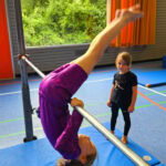 Ein Mädchen in pinkem Turnanzug ist gerade dabei einen Hüftaufzug an der Reckstange zu machen. Sie ist gerade in eine Kopfüber-Position. Dahinter steht ein Mädchen un schaut zu.