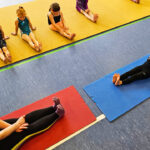 Eine Trainerin und ein Mädchen sitzen sich auf 2 Joga-Matten mit gestreckten Beinen gegenüber. Eine Gruppe Mödchen sitzt auf einem Bodenläufer und ahmt die Bewegungen nach.