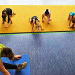 Eine Gruppe junger Mädchen kniet auf einem gelben Bodenläufer und dehnen sich für den Spagat. Links unten im Bild ist die Trainerin in der gleichen Pose zu sehen.