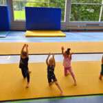 Eine Gruppe junger Mädchen kniet auf einem gelben Bodenläufer, ein Bein nach vorne ausgestreckt. Dabei strecken alle die Hände nach oben. Der Kopf und die ausgestreckten Arme der Trainerin sind links unten im Bild zu sehen.